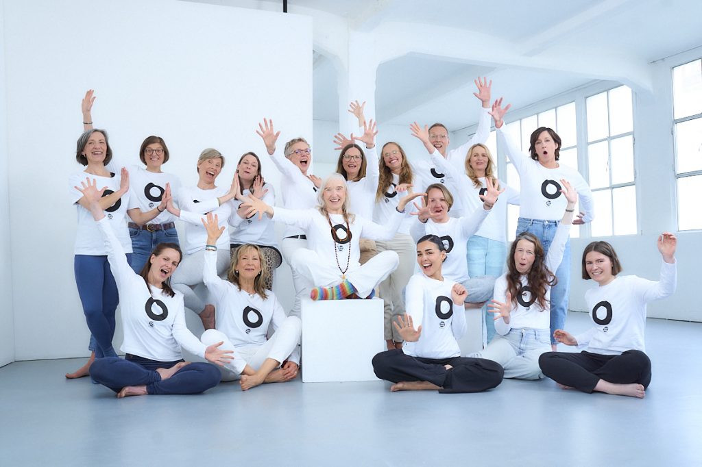 Ein heller Raum. Fröhliche Frauen, das Team von YOGAHILFT, alle in weißen T-Shirts mit schwarzem O.